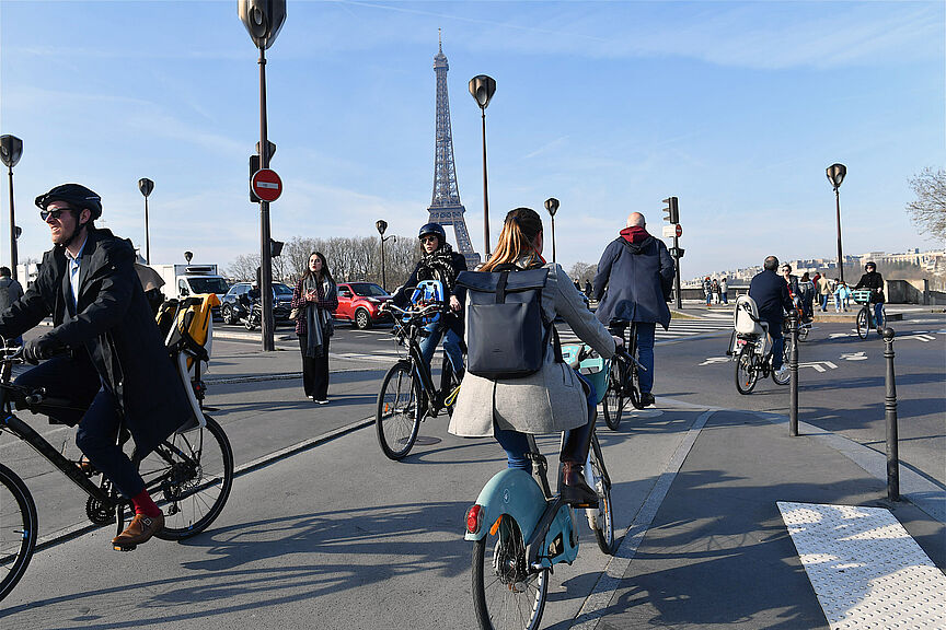 Radfahrende in Paris Menschen auf Fahrrädern überqueren eine Straße in Paris. Im Hintergrund ist der Eiffelturm zu sehen.